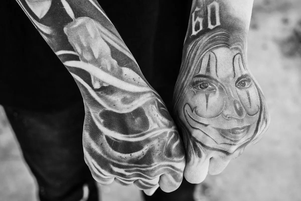 Black-and-grey payasa clown face and Old English numerals tattooed across the hands