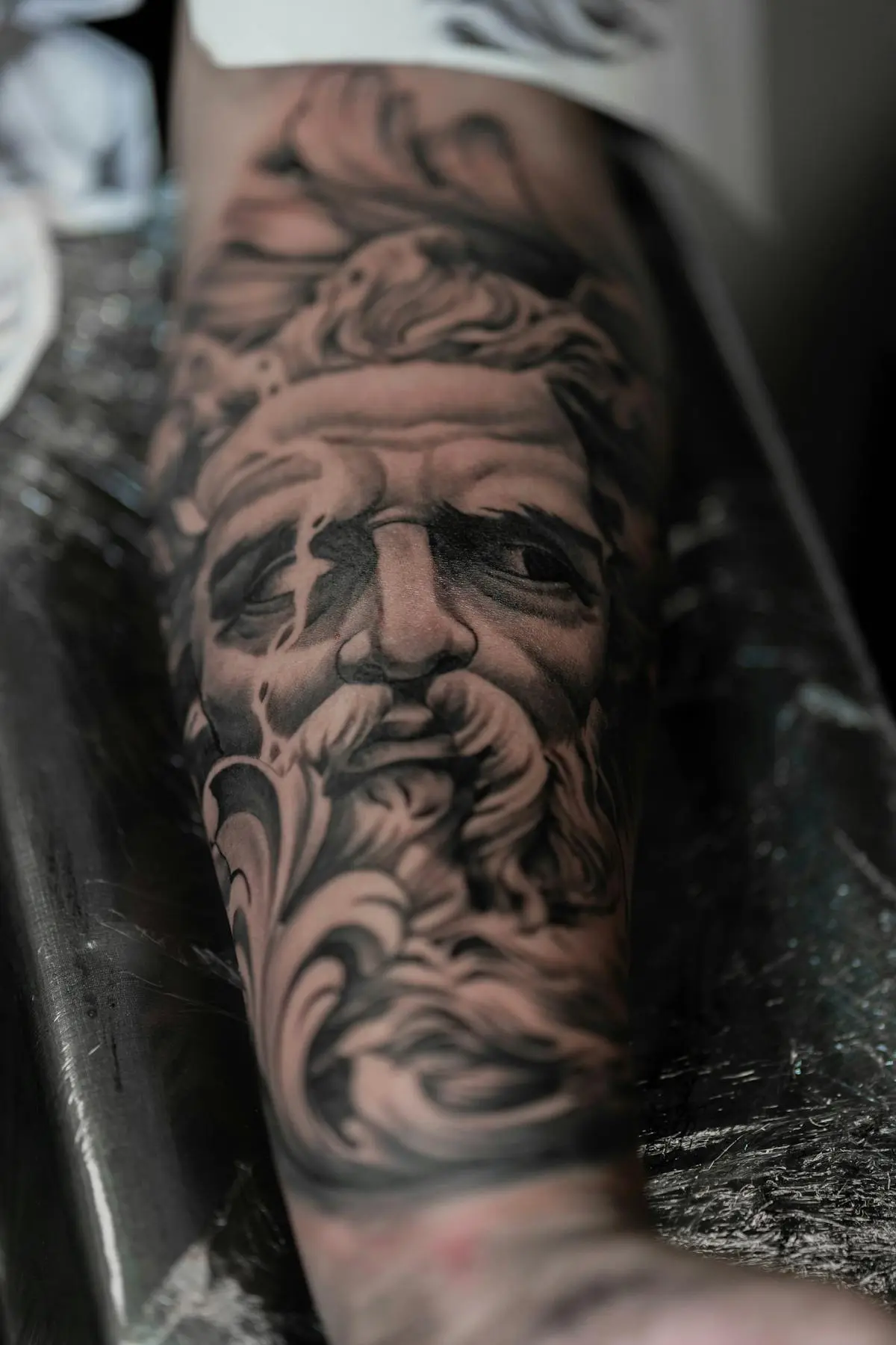 Black-and-grey photorealistic bearded statue portrait tattoo with filigree on the forearm