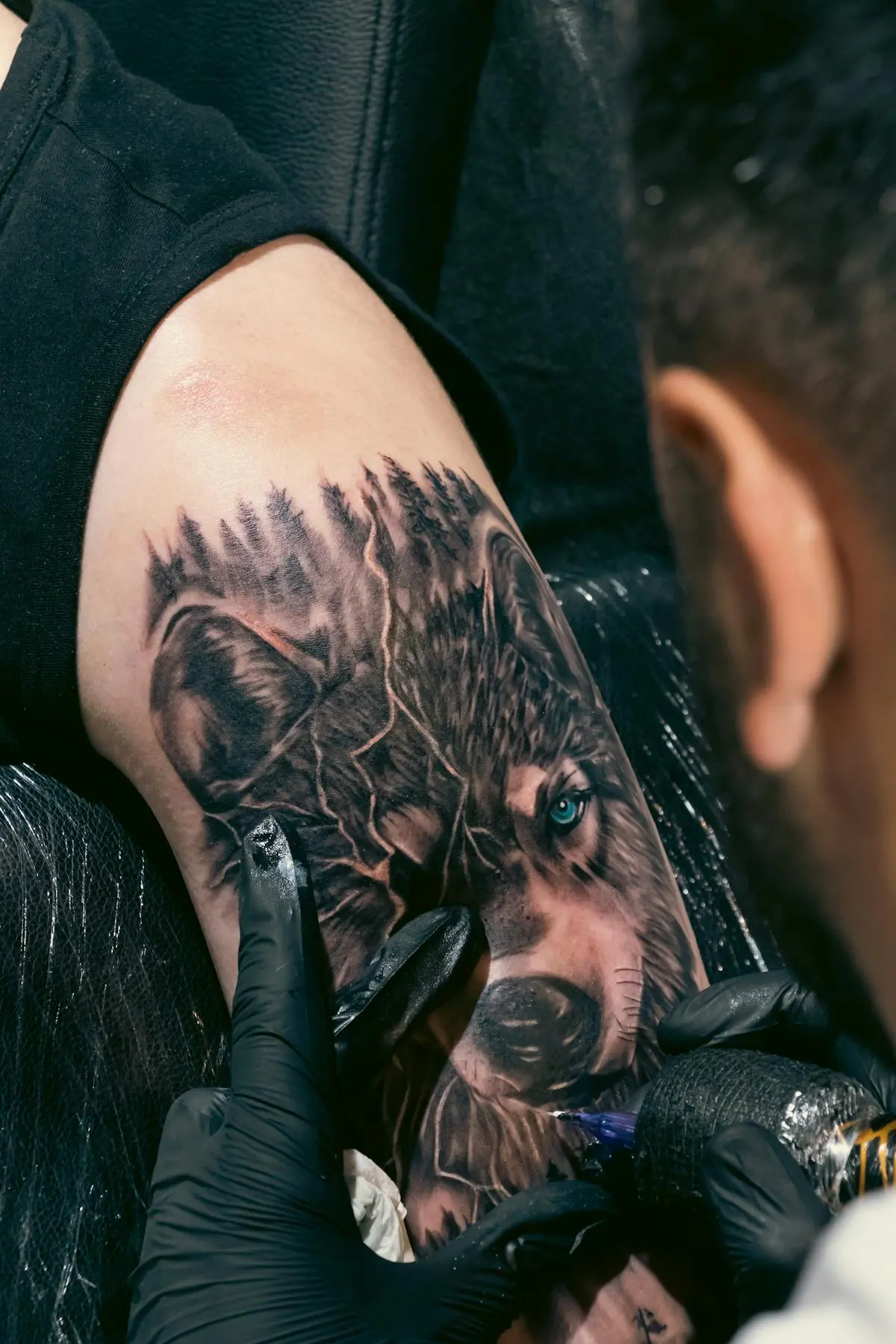 Realistic wolf head tattoo with a blue eye and forest detail being inked on the inner arm